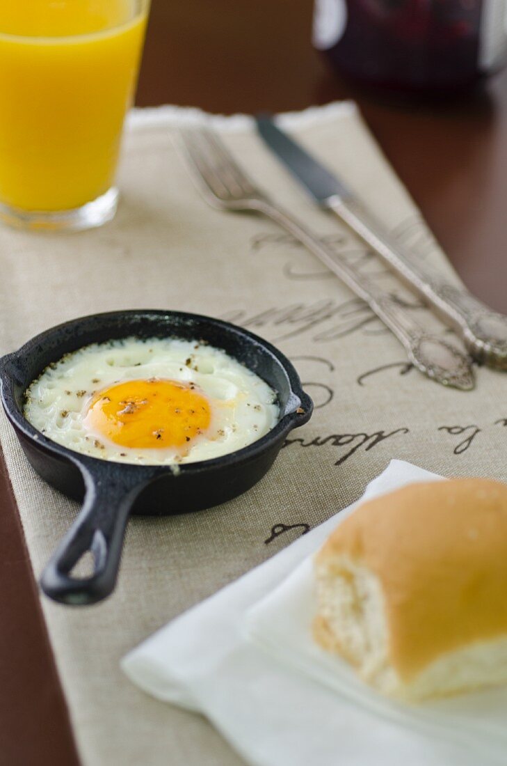 Fried egg in little frying pan with bread roll and orange juice