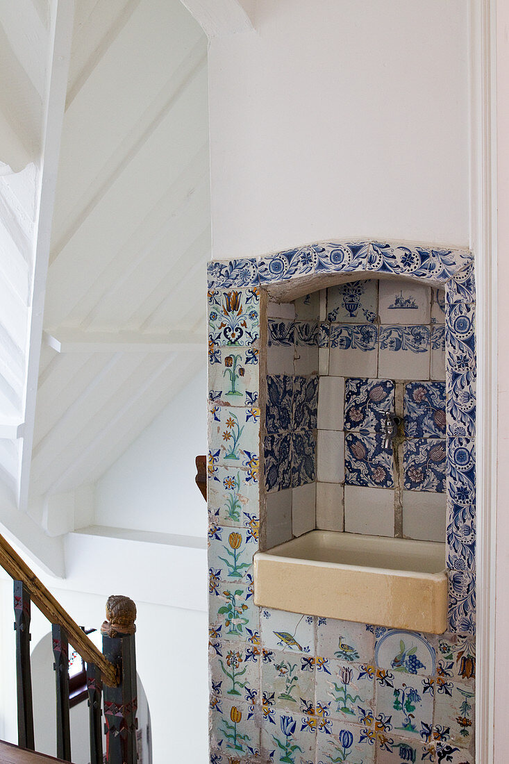 A hallway with an antique coloured wooden banister and an old wash basin in a wall niche with historic Delft tiles