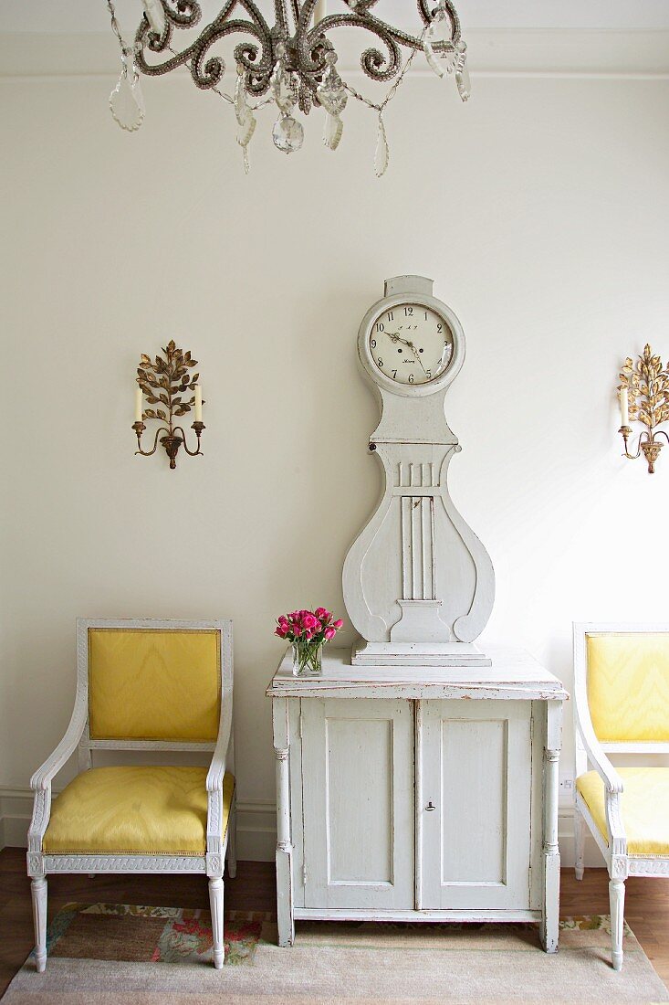 Baroque armchairs with yellow covers next to white-painted longcase clock on cabinet