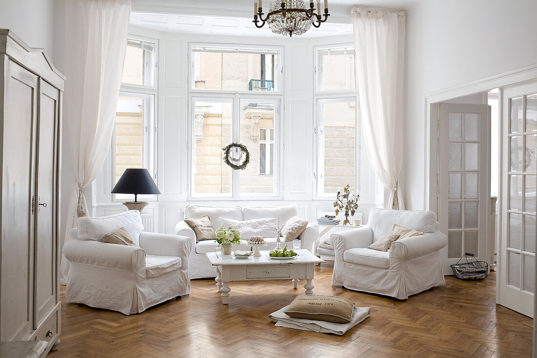 Sofa set with white loose covers and coffee table below chandelier in bay window of living room in period apartment