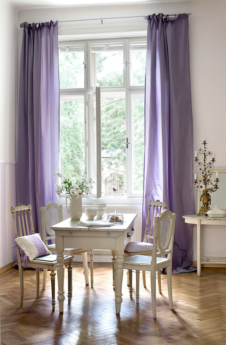 Old French chairs around wooden table in Gustavian style next to window with floor-length purple curtains