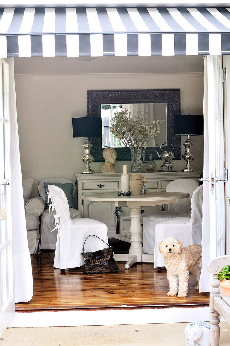 View from terrace with striped awning into white, country house living room with polished floorboards