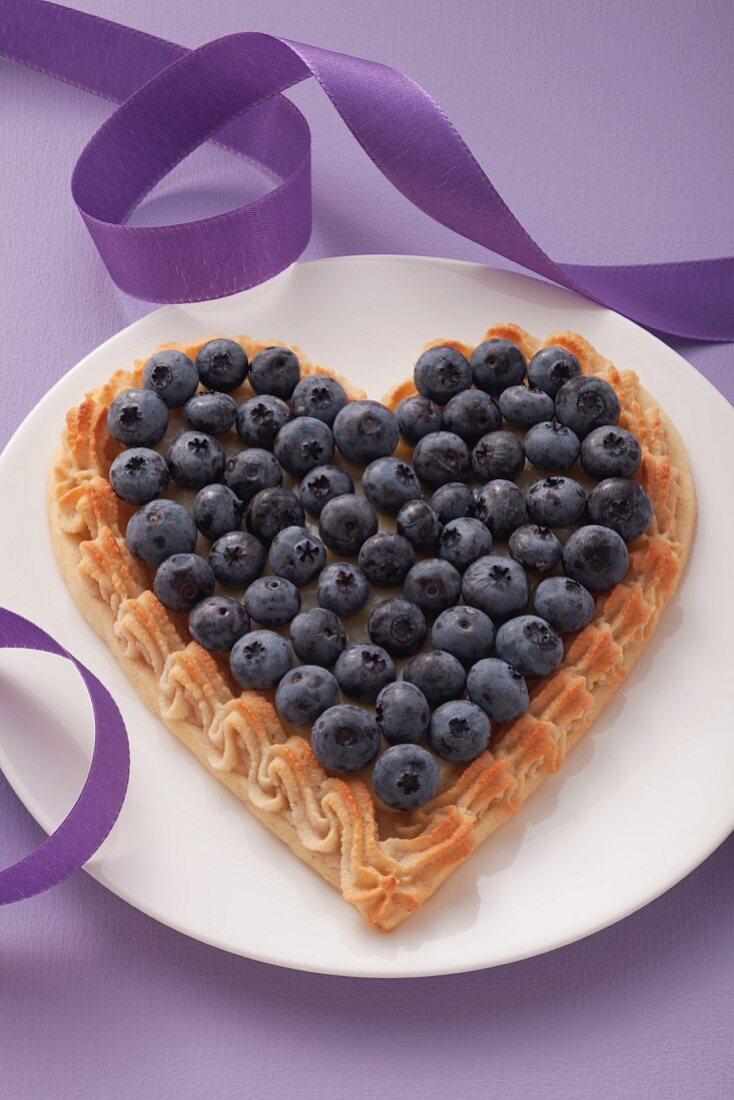 A heart-shaped blueberry tart on a plate with a purple ribbon