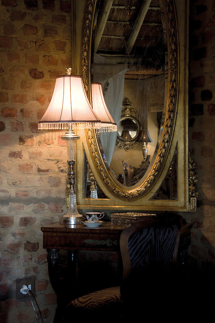 Tischlampe auf antikem Wandtisch vor Spiegel in Goldrahmen an Ziegelwand