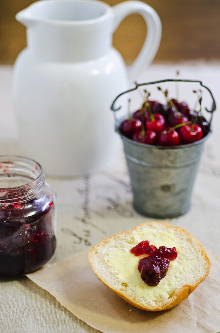 Half a bread roll topped with butter and cherry jam with a bucket of fresh cherries in the background