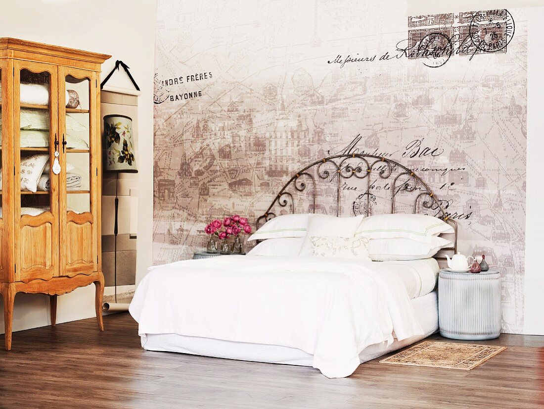 Bed with white linen and vintage metal frame against faded painting on wall and rustic, Baroque-style display cabinet