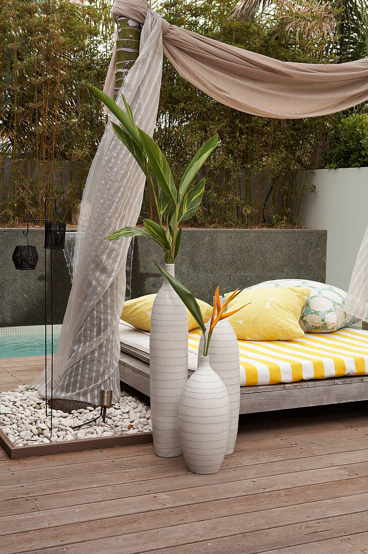 Vasenensemble auf Holzterrassenboden neben modernem Tagesbett mit Stoffhimmel an Palme befestigt