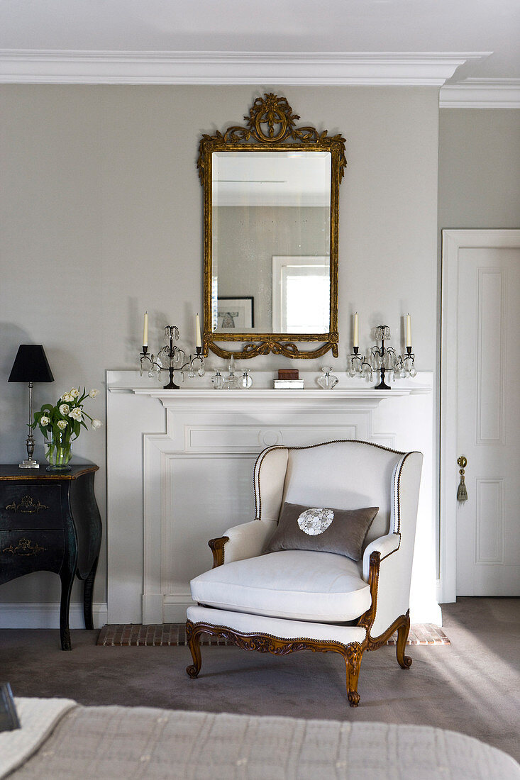 Elegant living room with armchair in front of disused fireplace, candelabras on mantelpiece and antique mirror