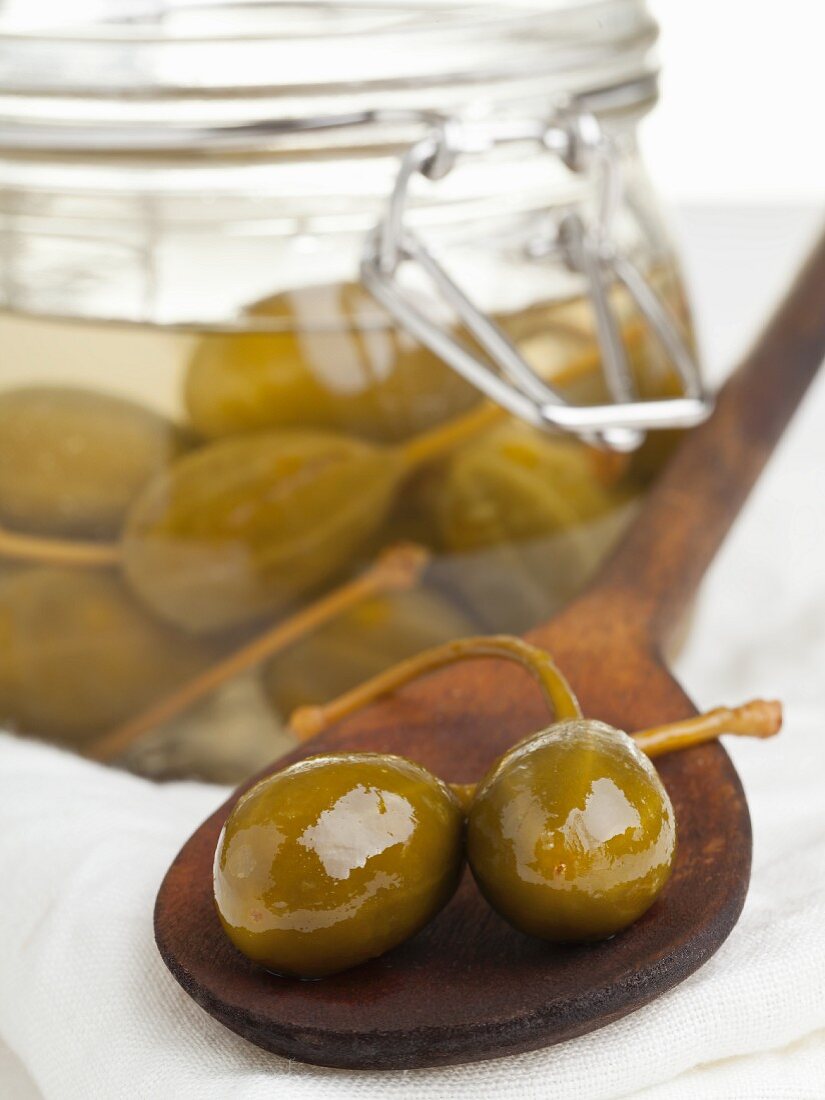 A jar of capers pickled in vinegar and some on a wooden spoon