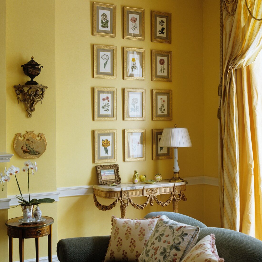 View across modern sofa to antique console table against yellow-painted wall and gilt-framed pictures with floral motifs