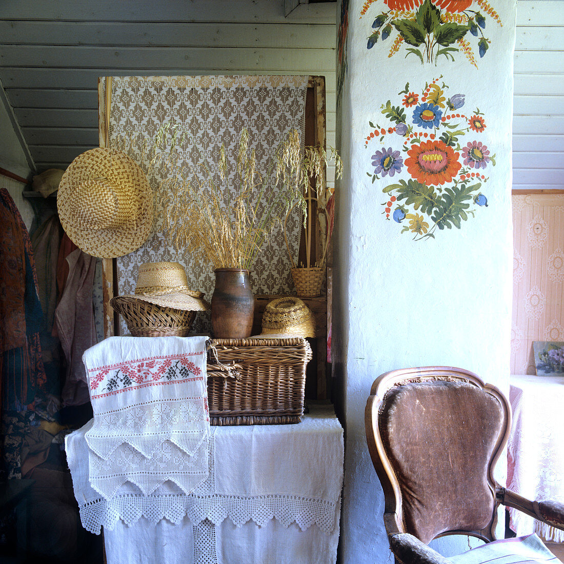 Ablage mit bestickter Leinenwäsche, Körben und Strohhüten; auf der Schornsteinsäule fröhliche Wandmalereien mit einfachen Blumenmotiven im bäuerlichen Stil