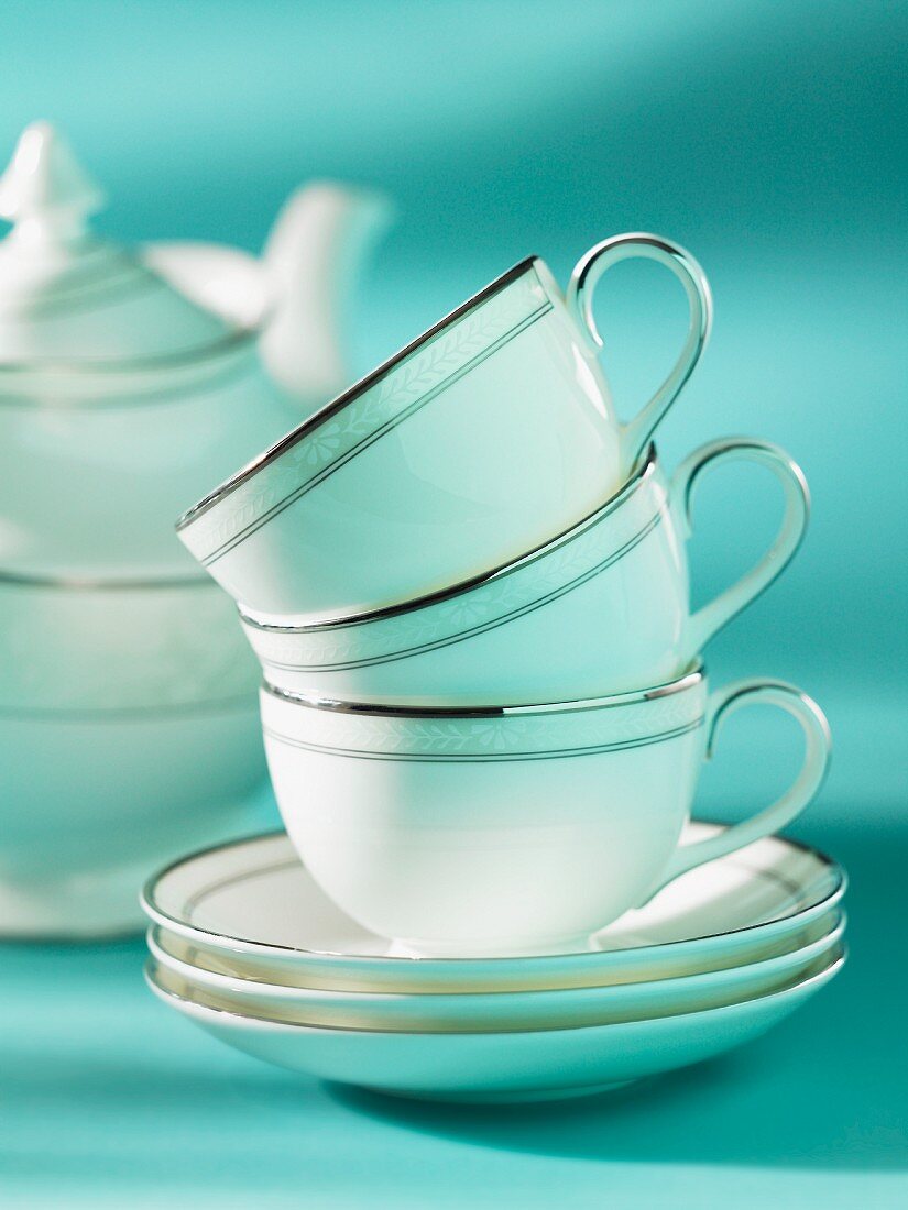 A stack of tea cups on a stack of saucers with a teapot in the background