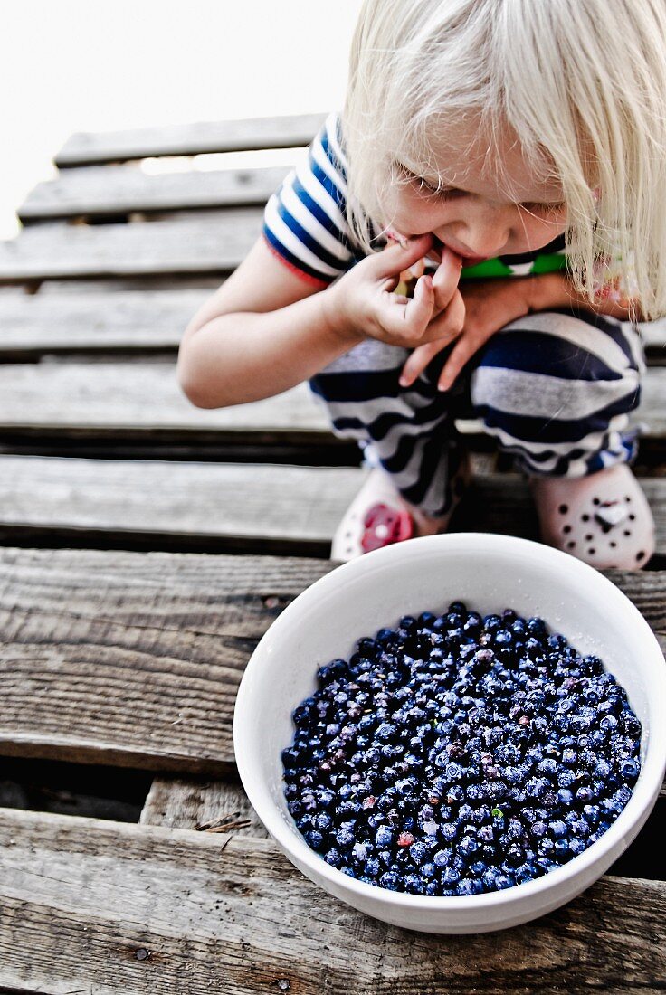 A child crouching in front of a bowl of fresh blueberries