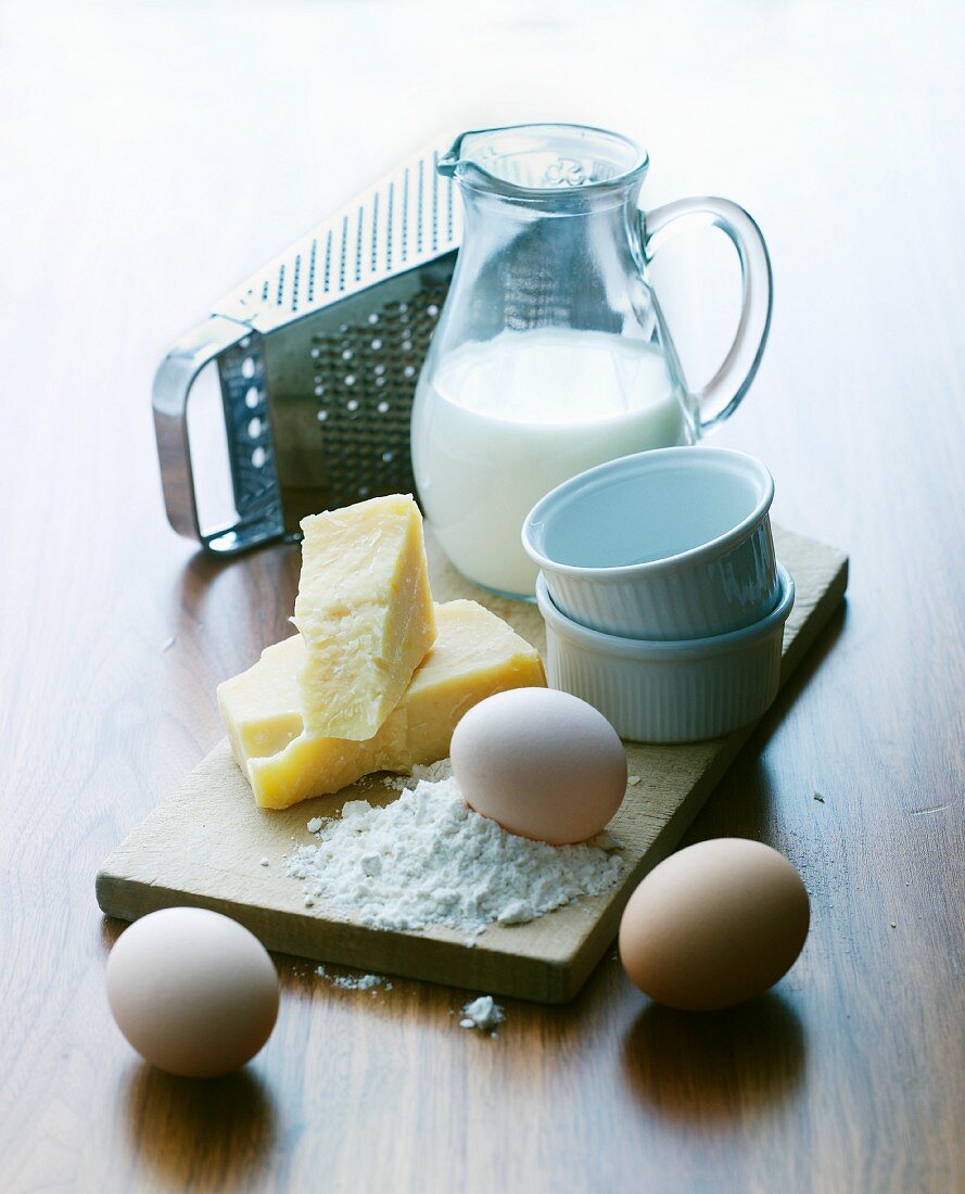 An arrangement of souffle ramekins, eggs, cheese, a cheese grater and milk