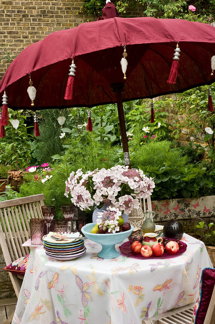 Hydrangeas, fruit, stacked plates and glasses on garden table below parasol