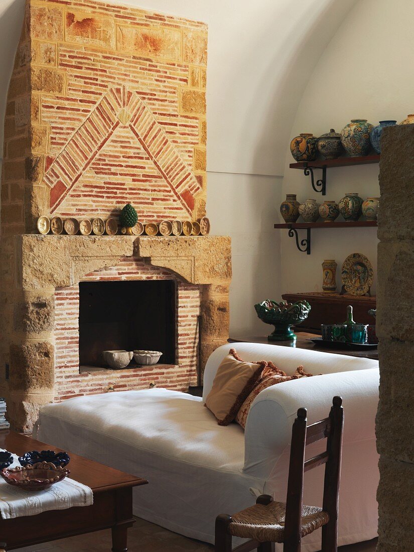 weiße Recamiere vor offenem Kamin mit antiker Ziegelmauer neben Wand mit Keramikvasen