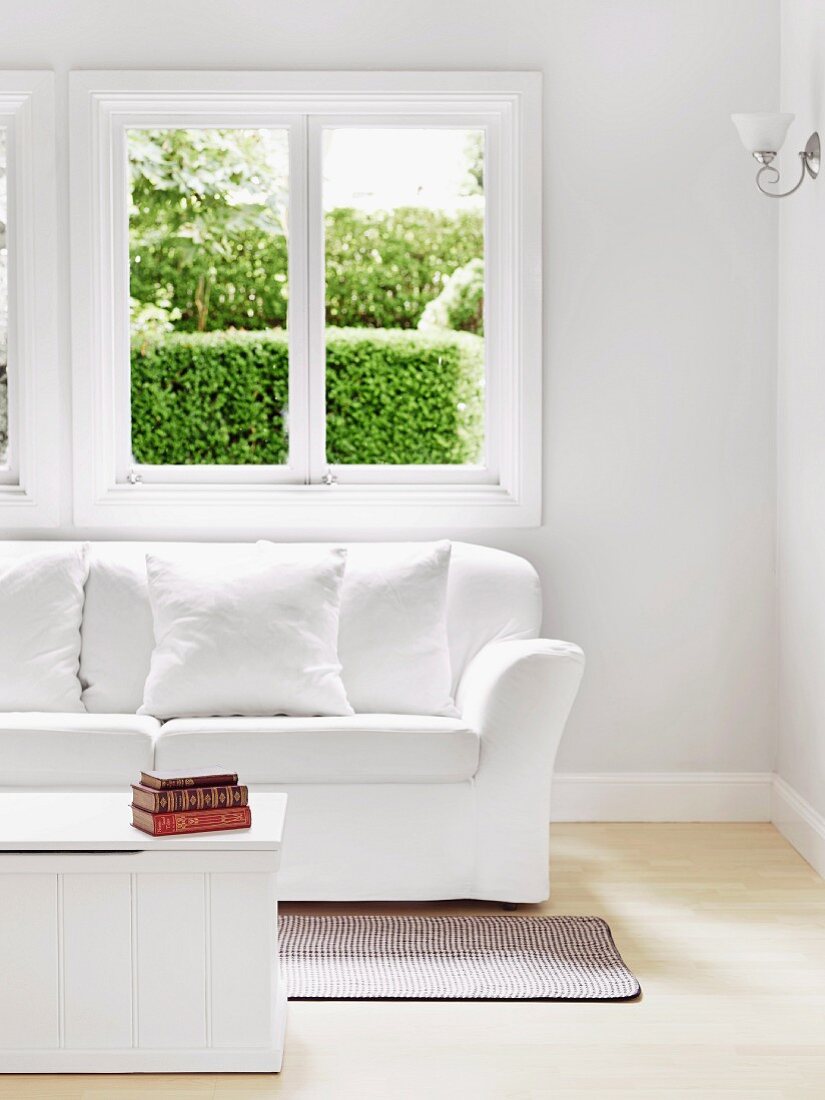 Simple, white wooden coffee table and sofa with white upholstery below window with view of garden