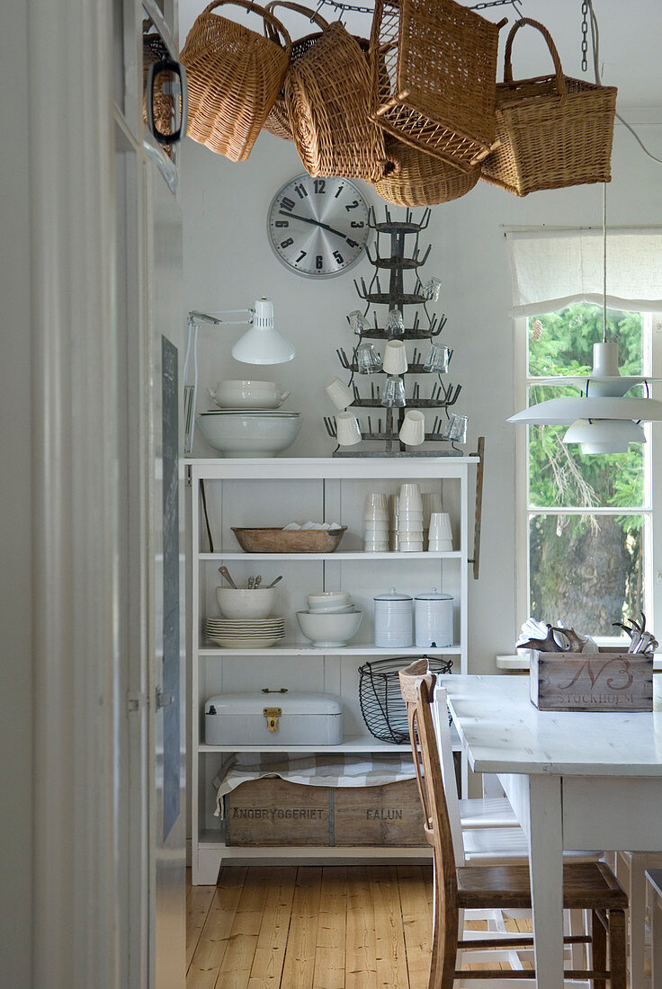 Corner of dining room with white dining set, baskets hanging from ceiling and white-painted, open-fronted shelves of crockery against wall