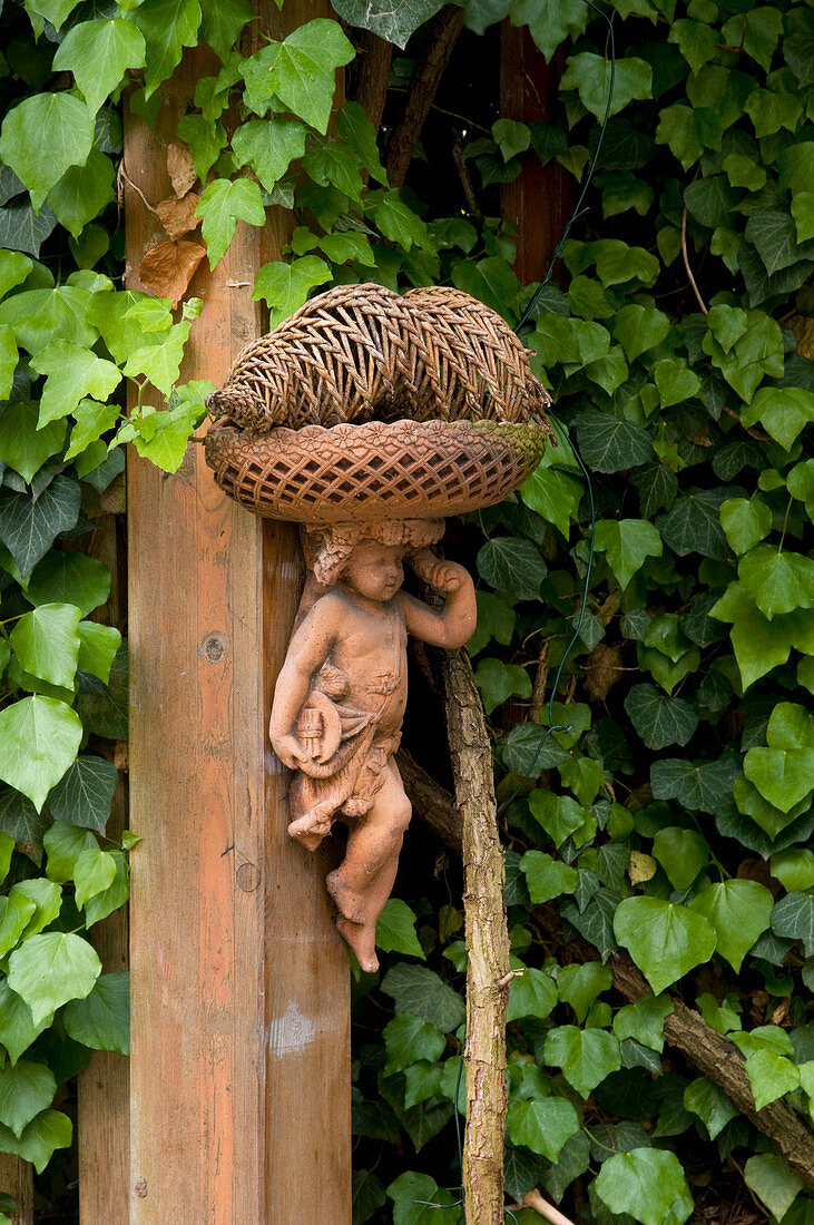 Terracotta figurine mounted on wooden post in front of ivy-grown hedge