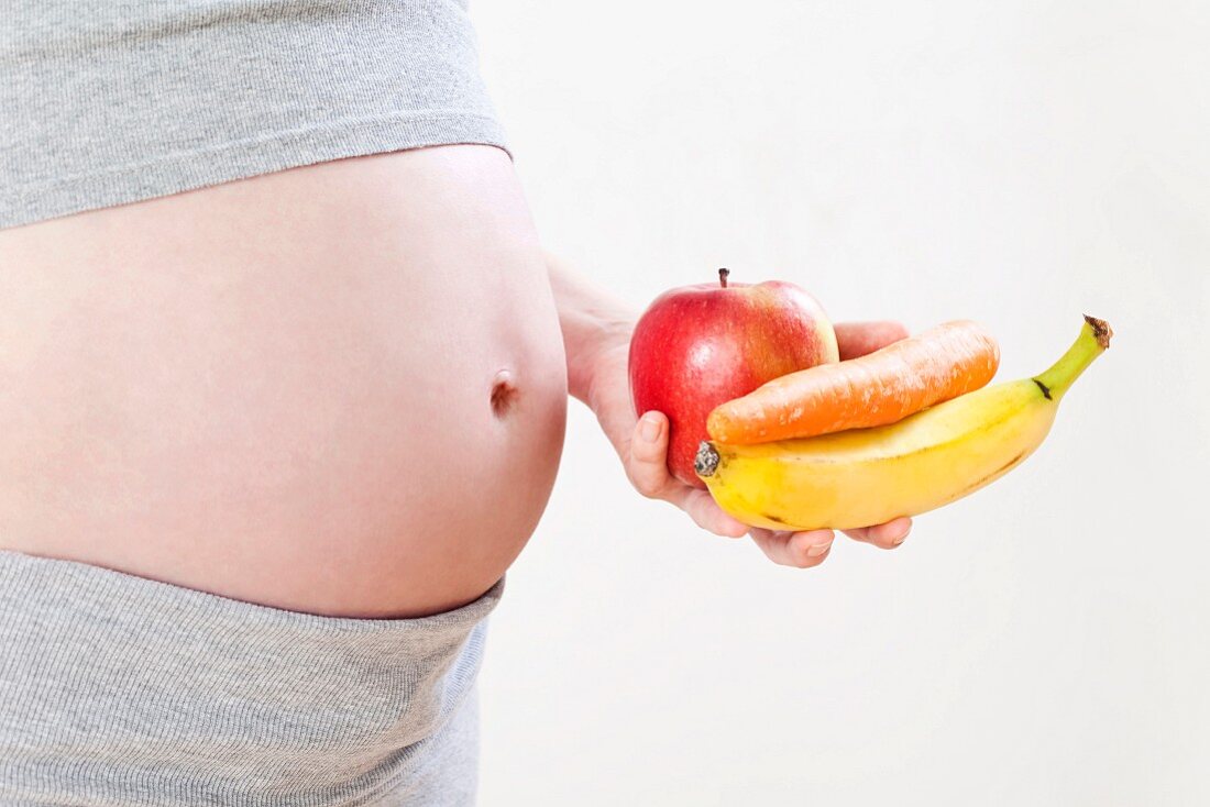 A pregnant woman holding an apple, a banana and a carrot