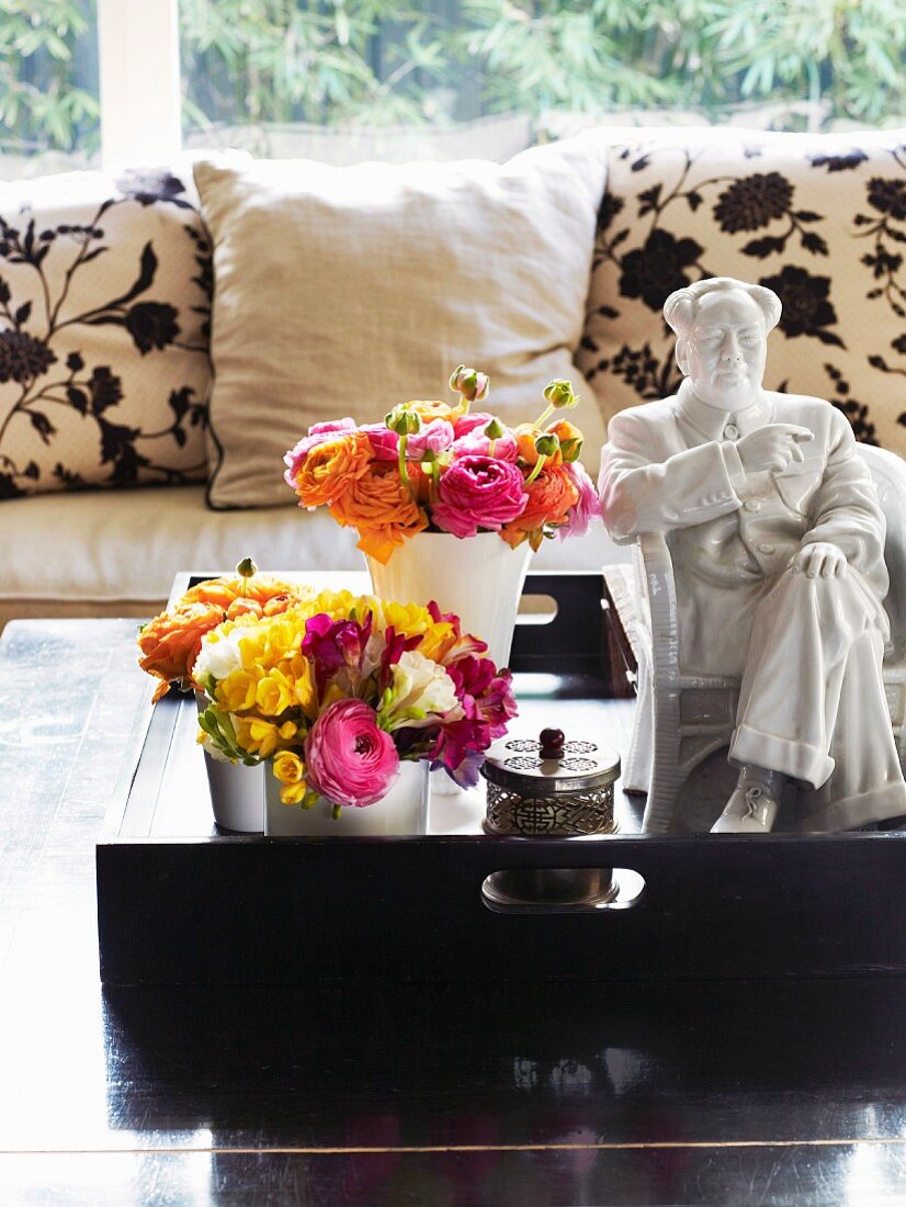 Bouquets in vases next to white china ornament on tray and coffee table in front of sofa with floral cushions