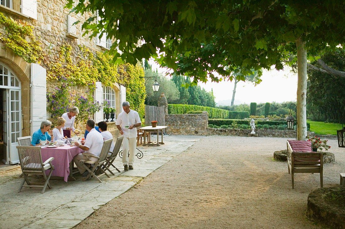 Guests taking breakfast on spacious terrace next to country house with view of Mediterranean garden