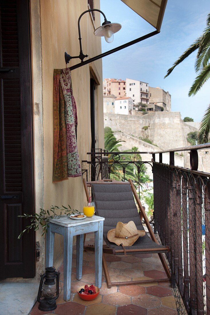 Simple, pale blue side table next to wooden deckchair with grey seat on small balcony of Mediterranean house with view of town on hillside