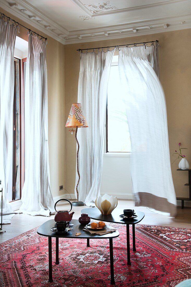 Herrschaftlicher Wohnraum mit Beistelltischen auf rotem Orientteppich und moderne Stehleuchte vor Fenster mit wehendem, bodenlangem Vorhang