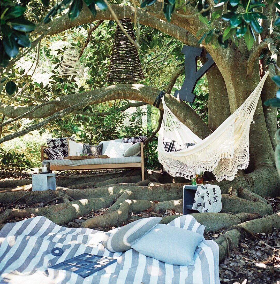 Gartenecke zum Ausruhen unter altem Baum mit Sitzbank, Hängematte und Decke zwischen Baumwurzeln