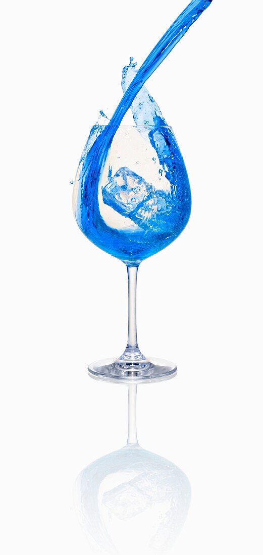 A blue cocktail being poured into a glass with ice cubes
