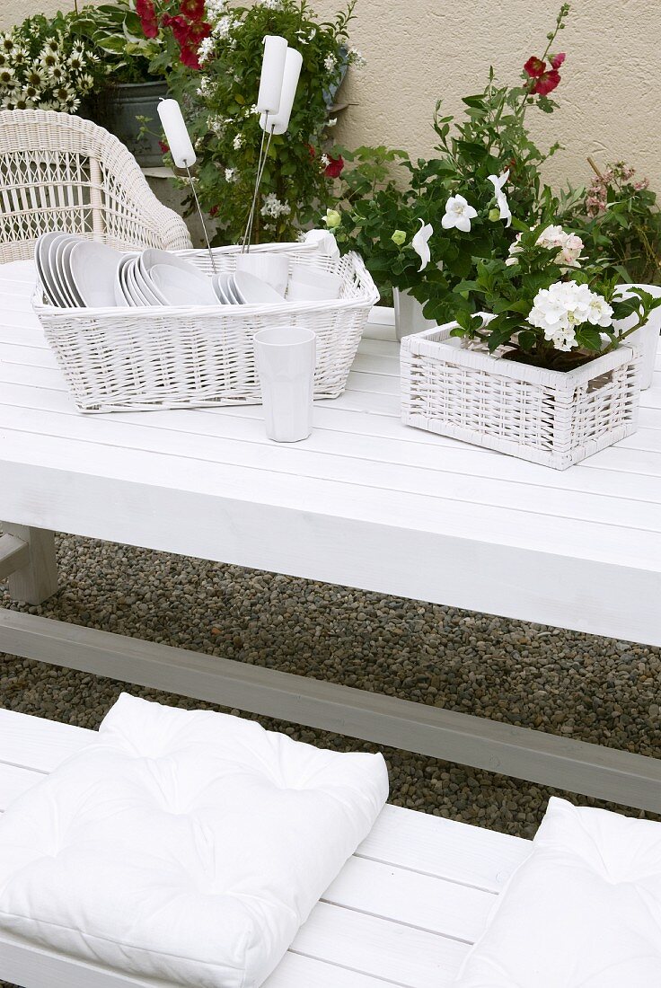 Crockery and potted flowering plants in wicker baskets on white garden table with matching bench