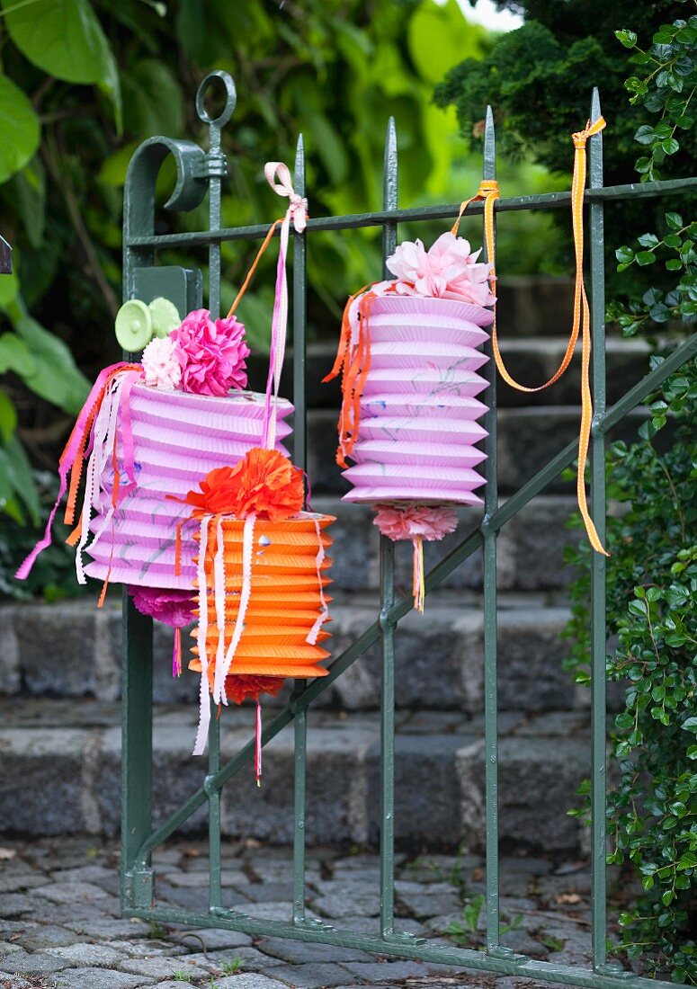 Concertina lanterns hanging on garden gate and decorated with ribbons, paper flowers and strips of paper