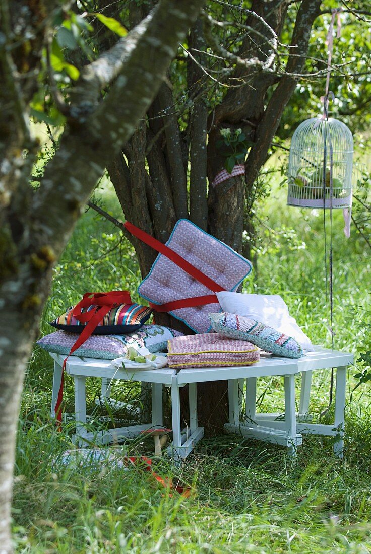 Sitzbank mit bunten Kissen um einen Baum herum und ein Vogelkäfig mit Äpfeln