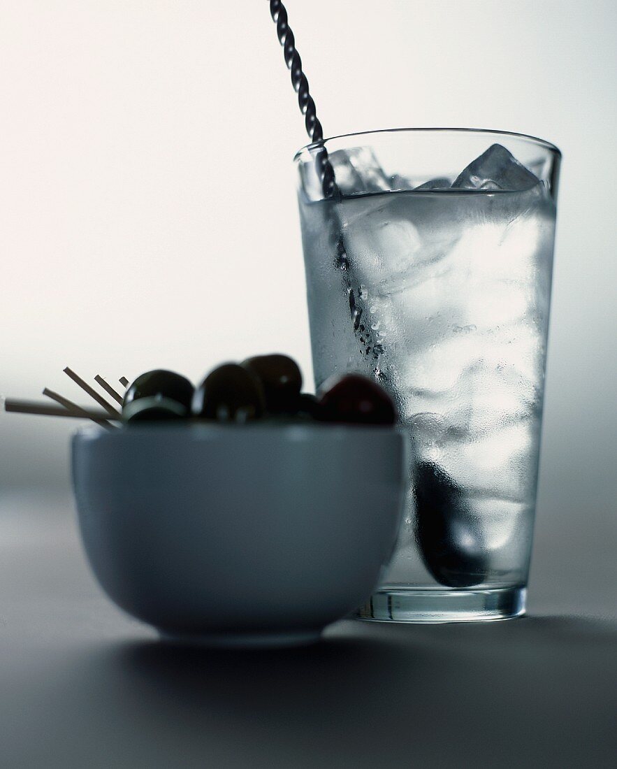 A cocktail with ice cubes and a bowl of olives