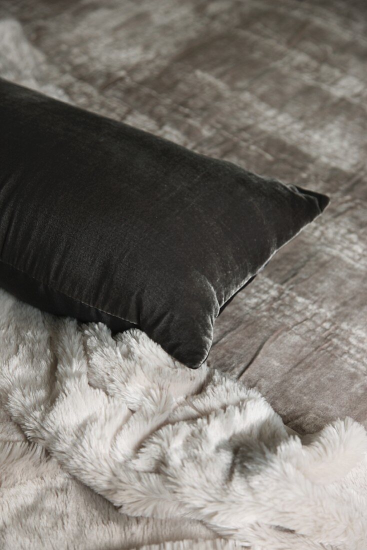 Kissen mit grauem Samtbezug neben Fell-Tagesdecke auf Bett