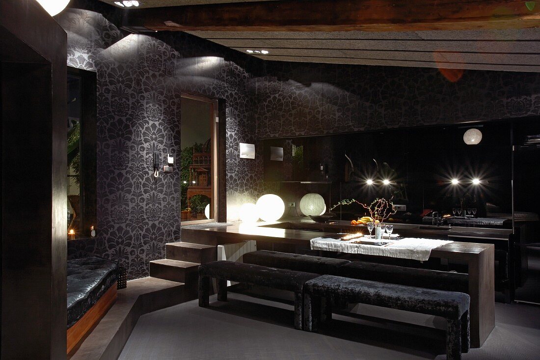 Minimalist dining table and upholstered benches in front of open-plan kitchen in interior in dark tones