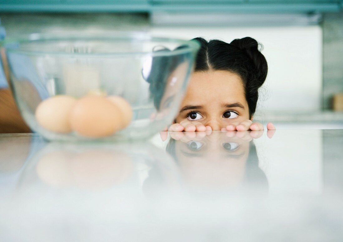 Mädchen schaut hinter dem Küchentisch hervor auf Schüssel mit Eiern