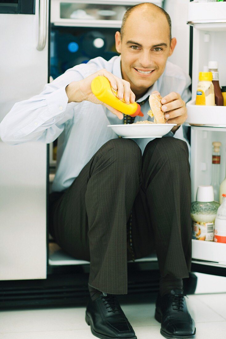 Man sitting in front of open refrigerator, putting mustard on hamburger