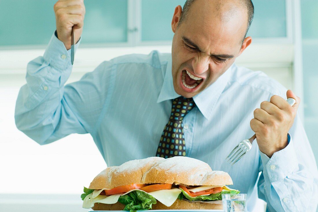 Man yelling and attacking large sandwich with knife and fork