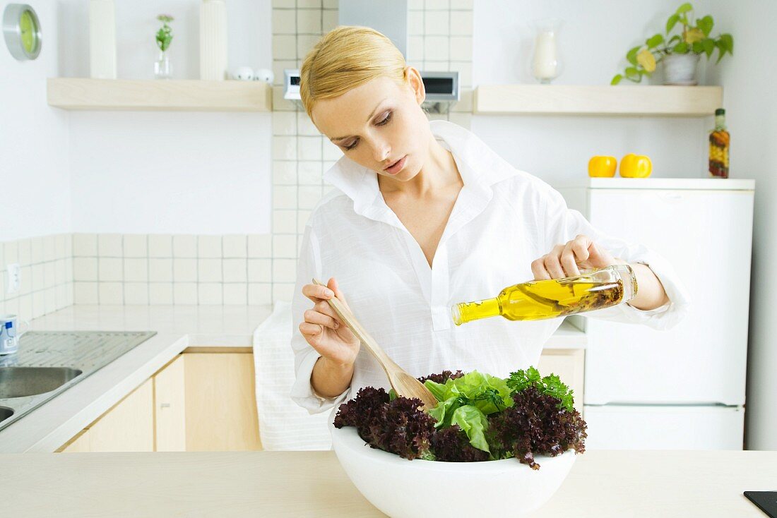 Woman standing in kitchen, pouring olive oil over salad