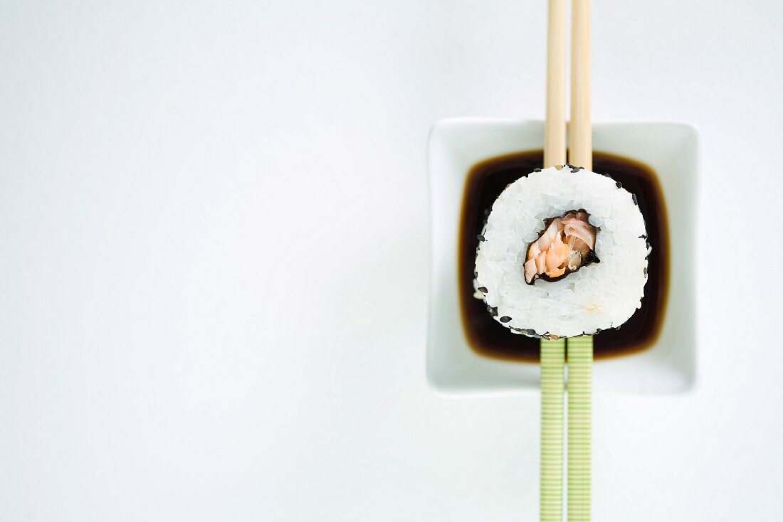 Single piece of maki sushi resting on chopsticks over sauce, overhead view