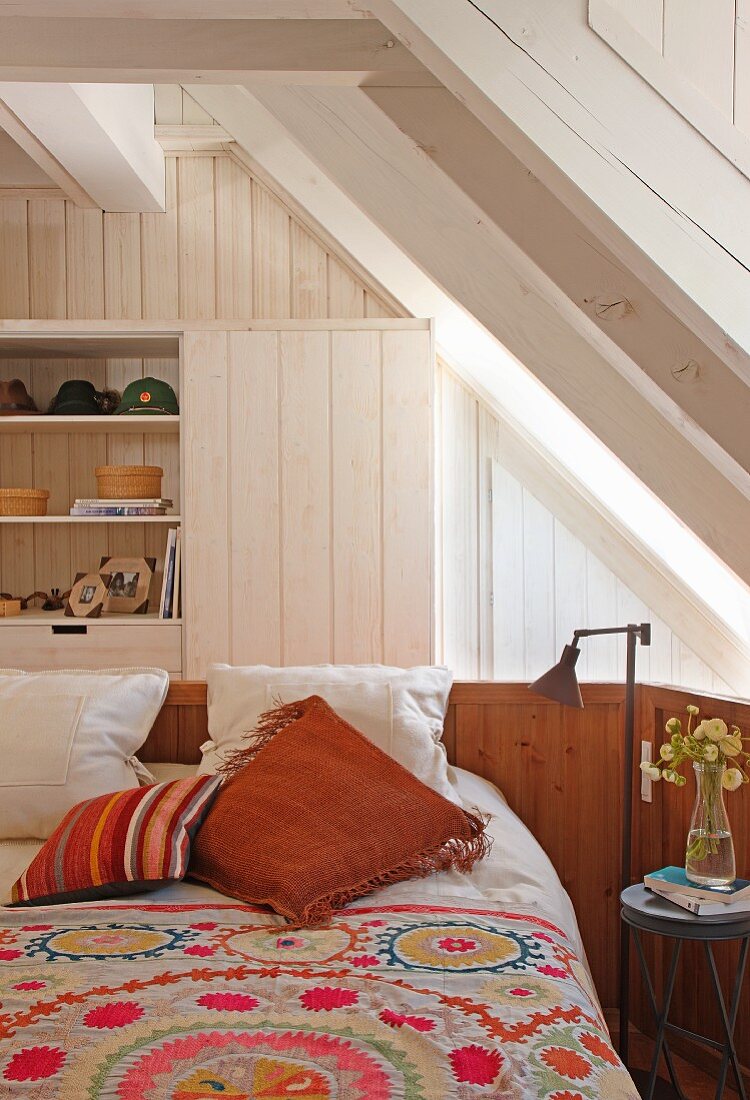 Bed with patterned bedspread in front of fitted cupboards below wood-clad sloping ceiling