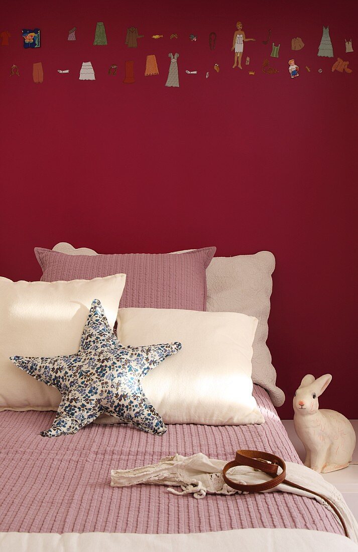 In Creme und Altrose dekoriertes Bett mit Seesternkissen und Hasenfigur vor dunkelrot getönter Wand mit einem Streifen aufgemalter Miniaturen