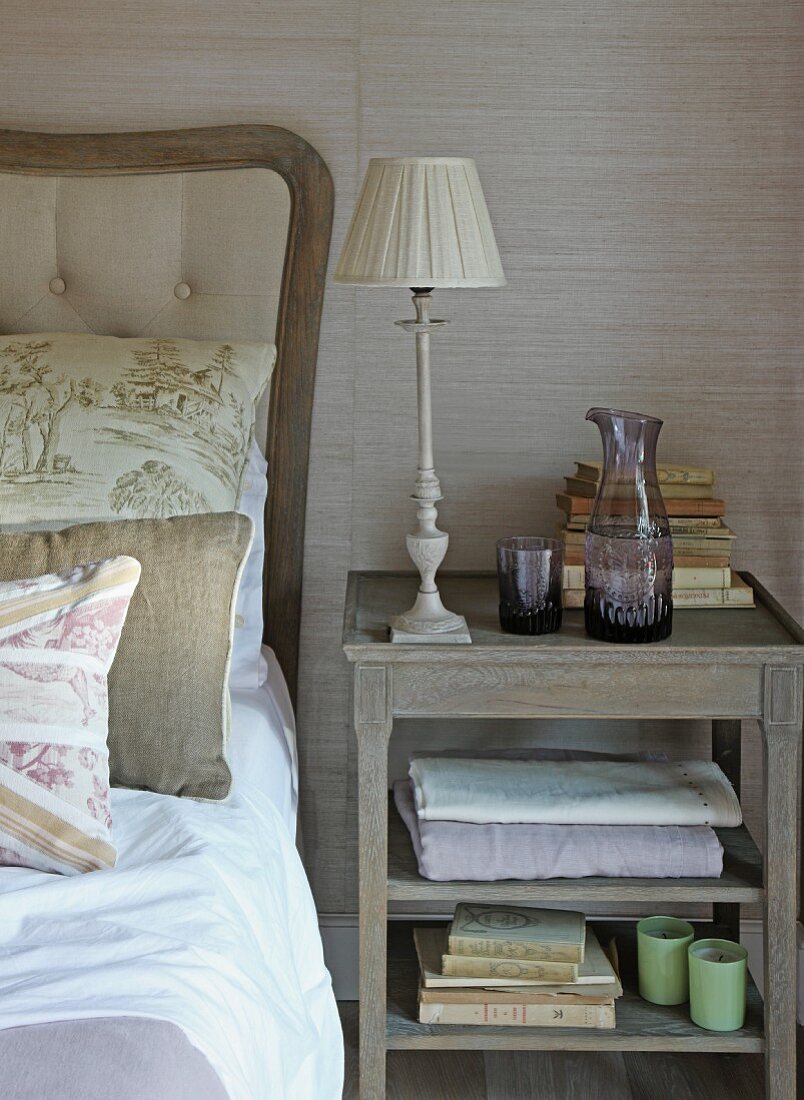 Table lamp and carafe of water on antique bedside table next to bed against grasscloth wallpaper