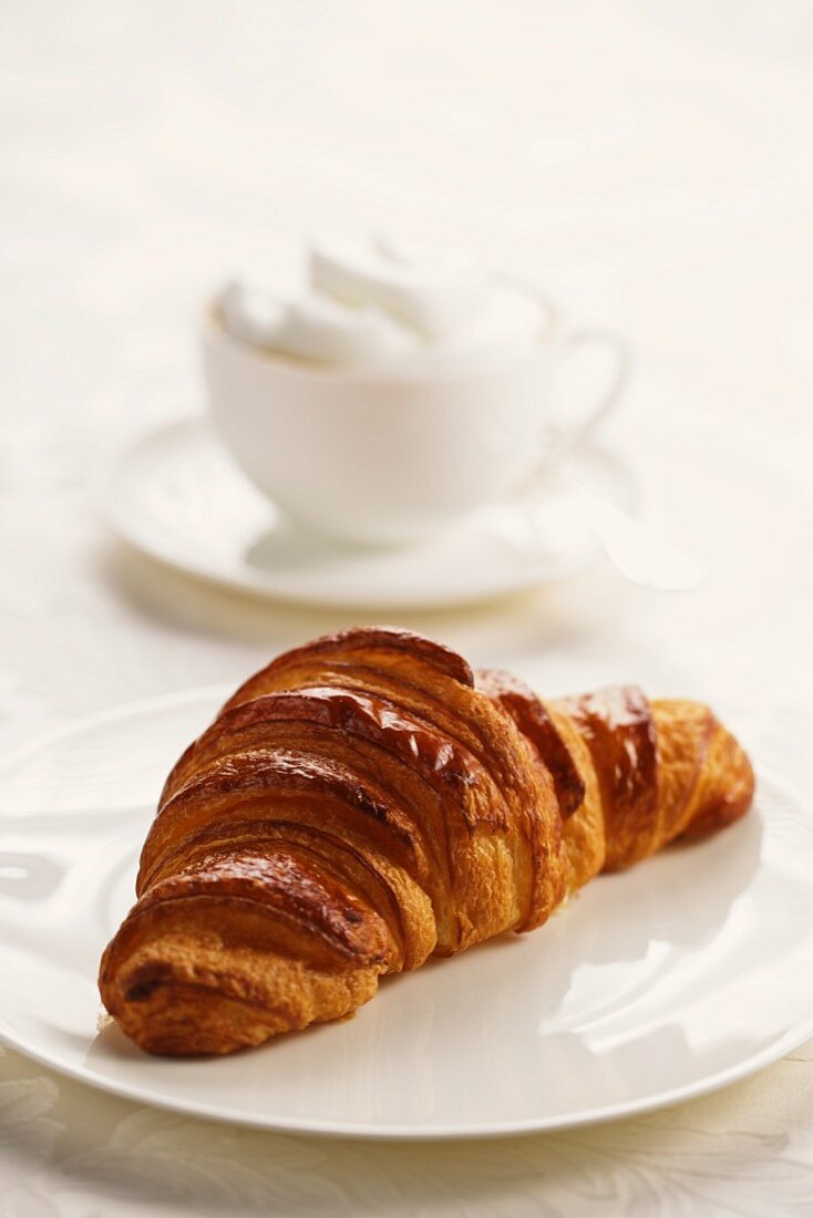 Ein Croissant auf Teller, im Hintergrund ein Cappuccino