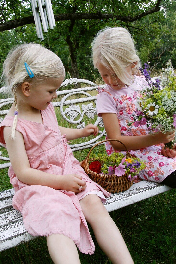 Zwei blonde Mädchen mit Blumen auf Gartenbank