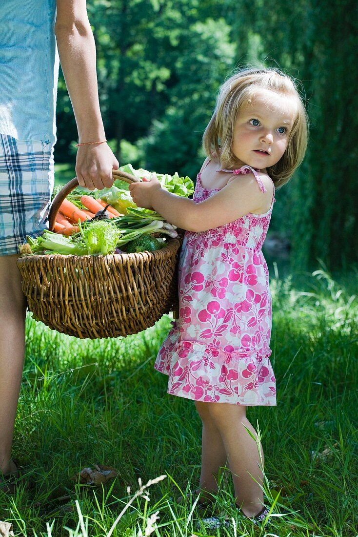 Little girl standing beside older sibling, holding large basket full of produce, cropped view