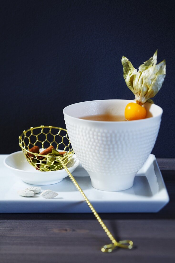 A cup of tea with a physalis and a tea strainer with a cinnamon stick and cardamom