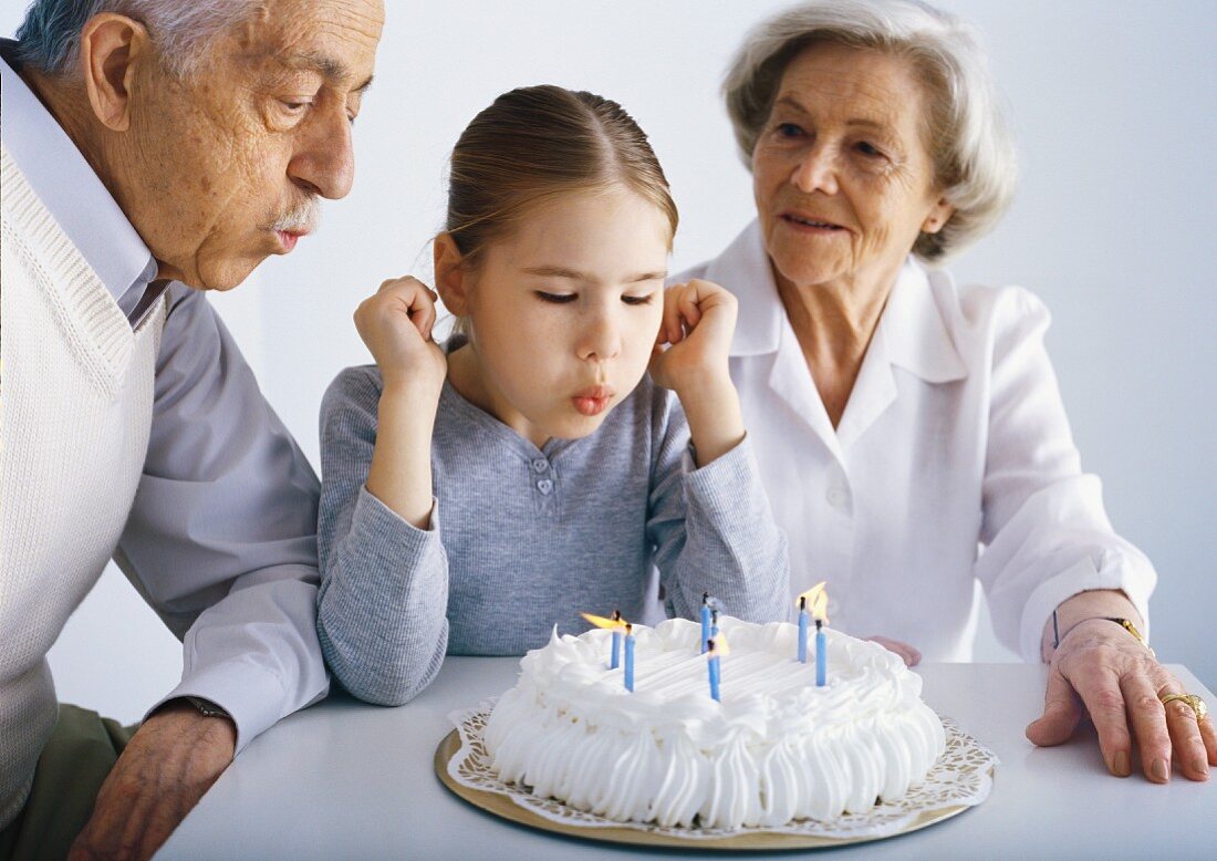 Grosseltern und Enkelin blasen Kerzen auf Geburtstagstorte aus