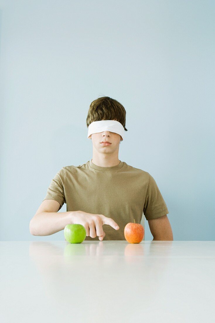 Young male wearing blindfold, pointing to one of two apples